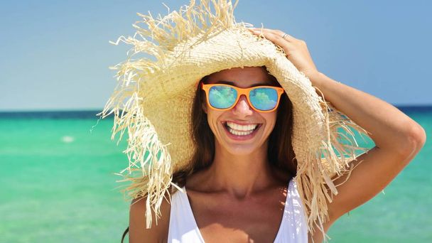 Beautiful happy woman at the sea, smiling in sunglasses, wearing a straw hat, in a white bathing suit, background of sea blue water. - Fotografie, Obrázek
