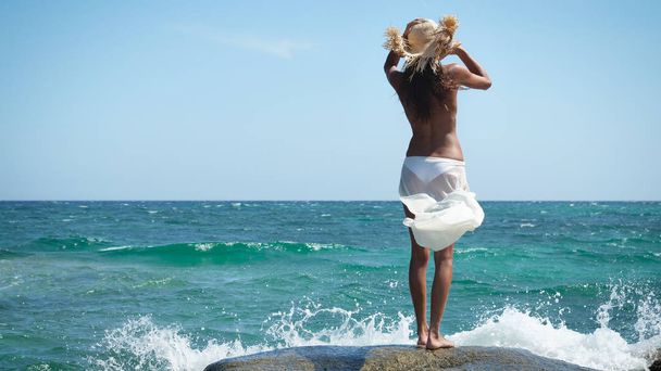 eine Frau steht auf den Felsen am Meer mit Strohhut im weißen Kleid, trägt Sonnenbrille, Hintergrund des blauen Meeres. - Foto, Bild