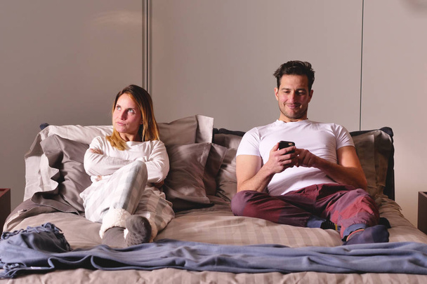 A married couple in bed use the phone separately to send messages or use social networks and no longer talk to each other. Concept of: new generations, separation, technology. - Zdjęcie, obraz