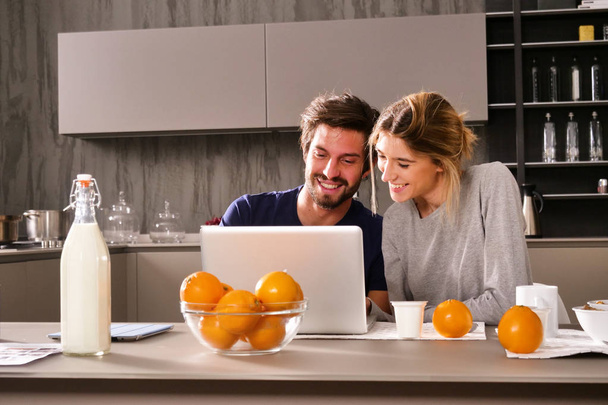 A couple in the kitchen looks at the computer with the souvenir photos of their holidays or of the past times while having breakfast and smiling happily. Concept of: family, technology, memories. - Φωτογραφία, εικόνα