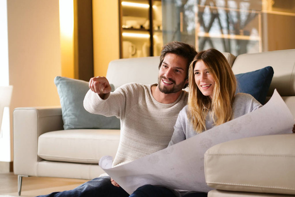 couple in love planning new home furniture while sitting on floor at sofa, man pointing finger  - 写真・画像