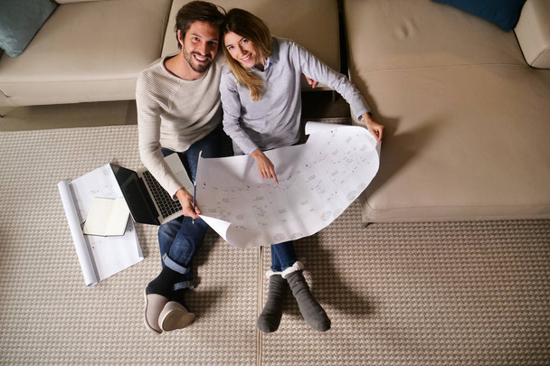 top view, couple in love planning new home furniture while sitting on floor at sofa and looking at camera - Zdjęcie, obraz