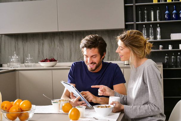 A couple in the kitchen looks at the tablet with the souvenir photos of their holidays or of the past times while having breakfast and smiling happily. Concept of: family, technology, memories. - Photo, image