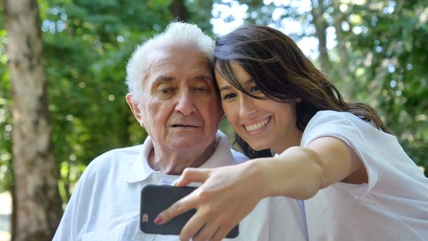 A nanny, a nurse, caring for the elderly, a girl (woman) and grandfather sit on a paperback and do selfie, in the park. Concept boarding house, sanatorium, house for the elderly, help for the elderly - Foto, Imagen