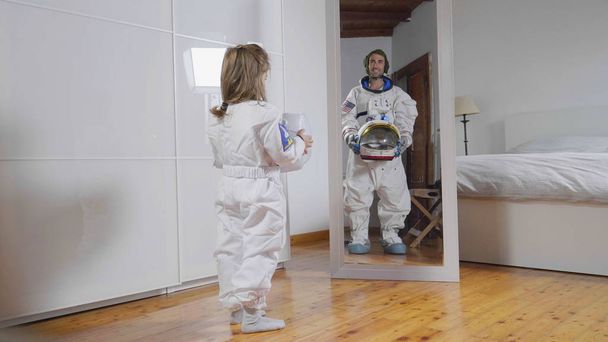 A child looks in front of the mirror wearing the astronaut's suit and looks great, his future, his job, his ambition to grow. Concept of: future, family, ambition, astronaut's work - Photo, Image
