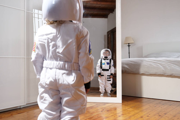 A child looks in front of the mirror wearing the astronaut's suit and looks great, his future, his job, his ambition to grow. Concept of: future, family, ambition, astronaut's work - Foto, immagini