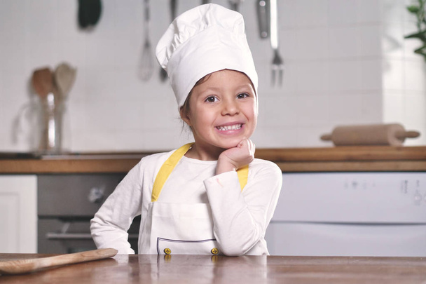 Portrait of a little girl in the kitchen dressed as a professional cook smiling and holding a traditional wooden kitchen spoon in her hand. Concept of: nutrition, cooking school, education and family. - Foto, Imagem