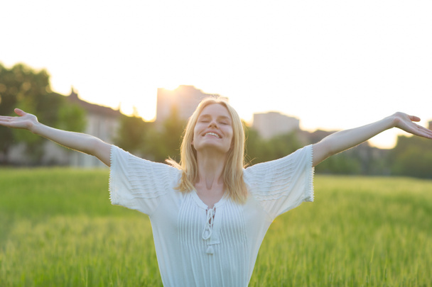 A happy beautiful woman enjoying the summer sunset in a green field. Arms in the air. - Photo, Image