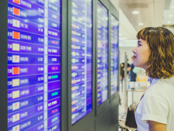 Asiatico donna capelli corti indossa camicia bianca controllo volo presso la scheda di informazioni di volo in aeroporto internazionale terminale
. - Foto, immagini