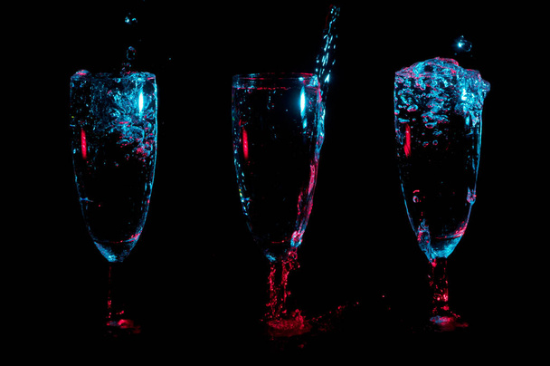 Una fila de tres copas de tulipán de champán con líquido transparente en luz de colores salpicando de ellos sobre un fondo negro
 - Foto, Imagen