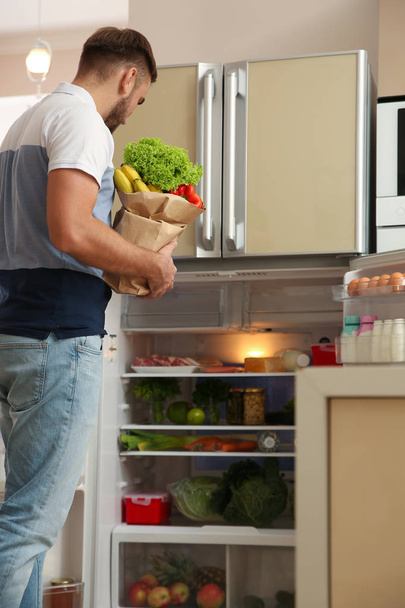 Young man with fresh food in paper bag near refrigerator at home - Photo, Image