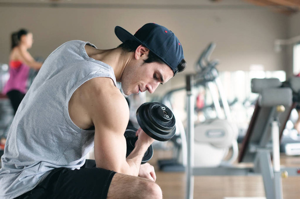 A guy, wearing a tank top in a gym, trains his body to stay fit and have defined muscles. The athlete raises heavy weights and fatigue. Concept of: sport, gym, muscles and fitness - Fotoğraf, Görsel