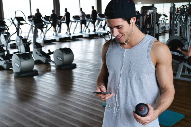 Portrait of a guy in a gym while resting between a workout and the other uses his phone to watch social networks or send messages. Concept of: sports, personal trainer, messages, telephone - Foto, imagen