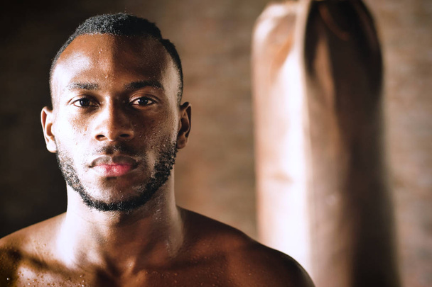 portrait of serious handsome shirtless muscular boxer african man posing on camera and standing at punching bag  - Foto, Imagem