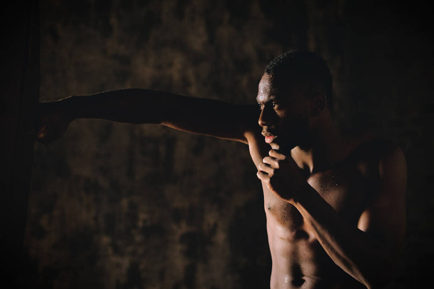 szép izmos afrikai félmeztelen férfi boxing boxzsák, sötét szobában  - Fotó, kép