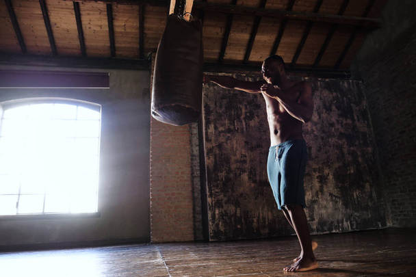 full length of handsome shirtless muscular african man boxing punching bag  - Фото, изображение