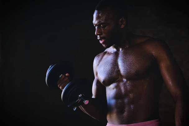 african athlete man with muscular biceps exercising with dumbbell in darkness  - Foto, afbeelding