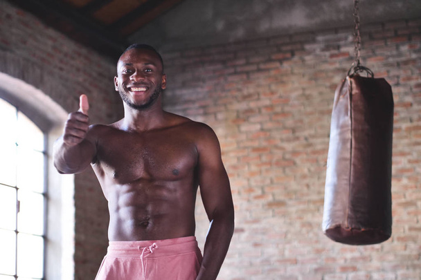 sonriente sin camisa musculoso boxeador africano hombre de pie en saco de boxeo y gestos pulgares hacia arriba
  - Foto, Imagen