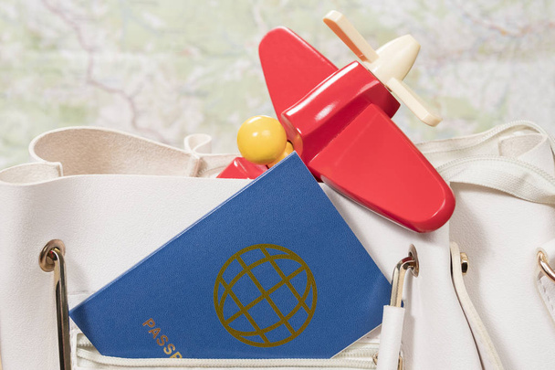passport and toy plane sticks out of a pocket of a white female bag, close-up, on a background of a map - Фото, изображение
