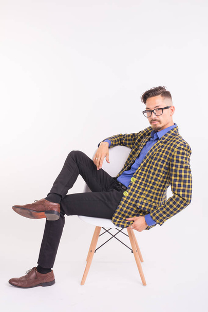 Concepto de moda y gente - hombre guapo barbudo seguro sentado en la silla sobre fondo blanco
 - Foto, Imagen