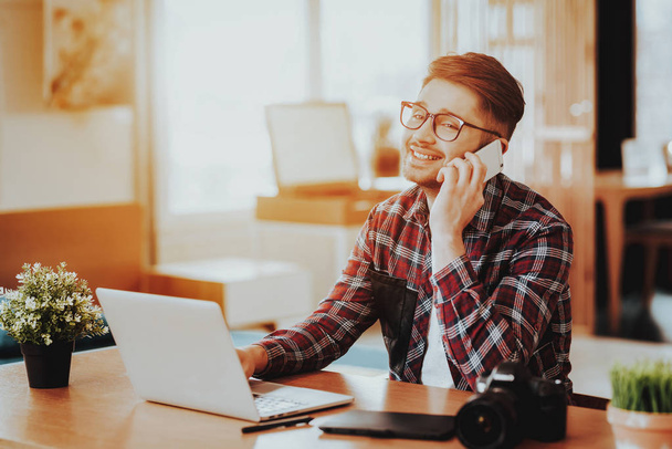 Happy Guy Talks Phone and Works on Laptop Remotely. Portrait of Happy Smiling Freelancer Wears Glasses Sits at Table Indoors. Distance Job and Freelancing Communication Concept - Foto, imagen