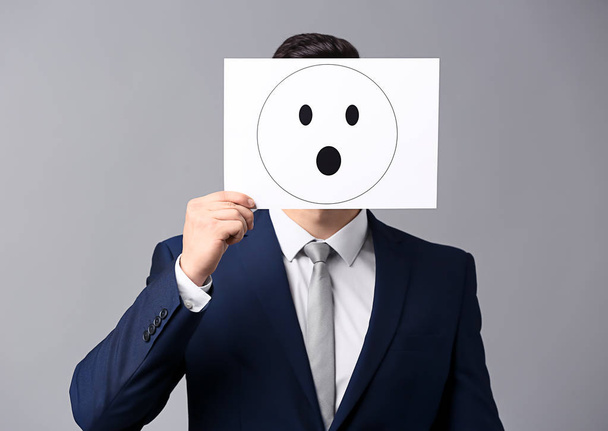 Businessman hiding face behind sheet of paper with drawn emoticon on grey background - Photo, Image