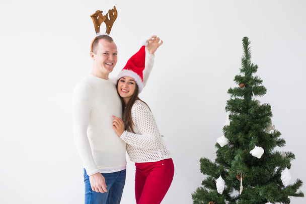 Funny loving couple near Christmas tree. Man wears deer horns and woman wears santa hat - Photo, Image