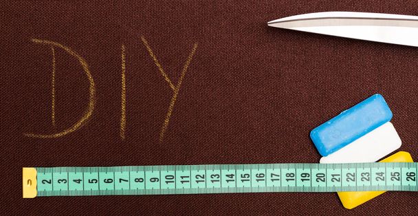 Brown fabric with DIY written on it underlined by measuring tape with pair of scissors and coloured tailoring chalk near as do it yourself hobby concept - Foto, Imagen