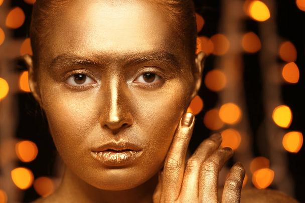 Beautiful young woman with golden paint on her body against defocused lights, closeup - Photo, Image