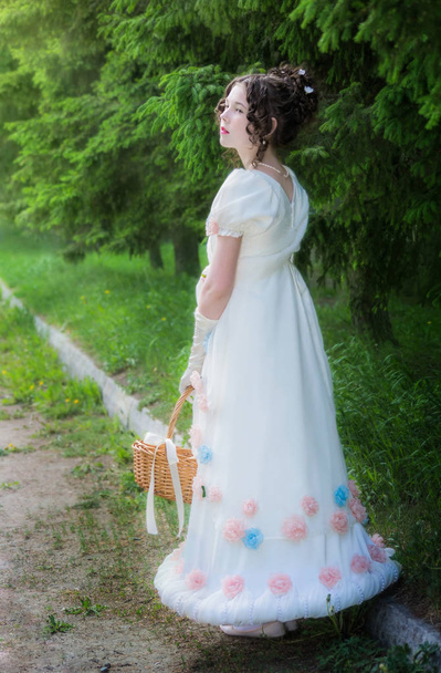 Beautiful girl in an historical bride dress with a wicker basket in her hands in a spring garden for a walk. - Foto, afbeelding