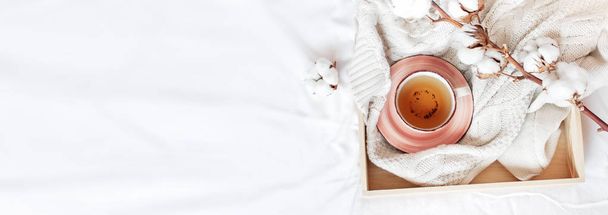 Cup of hot tea, wooden tray, branch of delicate cotton and knitted plaid sweater in bed. Cozy morning breakfast at home Lifestyle gentle female background Copy Space autumn winter concept - Photo, Image