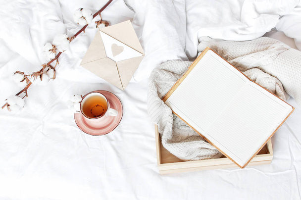 Cup of hot tea branch of cotton wooden tray knitted plaid sweater open notebook love letter in bed. Cozy morning breakfast at home Lifestyle gentle background Copy Space autumn winter concept - Photo, Image