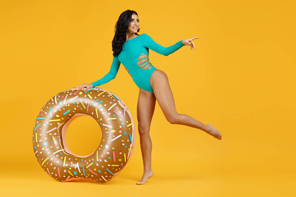 Full lenght portrait of an excited young fit woman dressed in swimsuit posing with inflatable ring and pointing finger away on the orange background. - 写真・画像