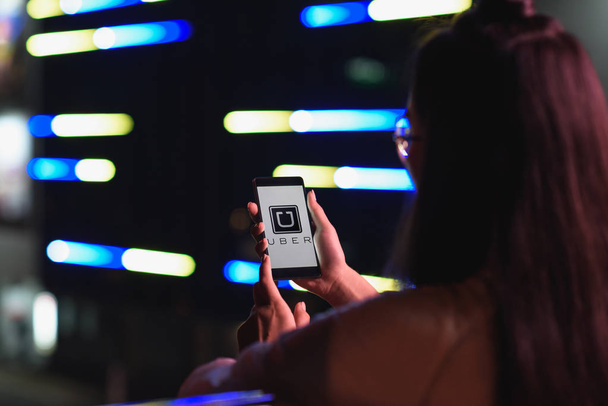 Achteraanzicht van meisje smartphone met geladen uber pagina houden op straat met neon licht in de avond, stad van de toekomst concept - Foto, afbeelding