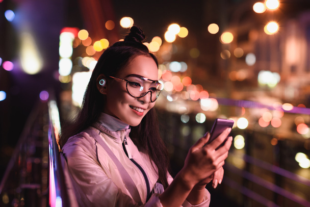 atractiva chica asiática con auriculares inalámbricos y gafas usando teléfono inteligente en la calle con luz de neón, ciudad del concepto futuro
 - Foto, Imagen