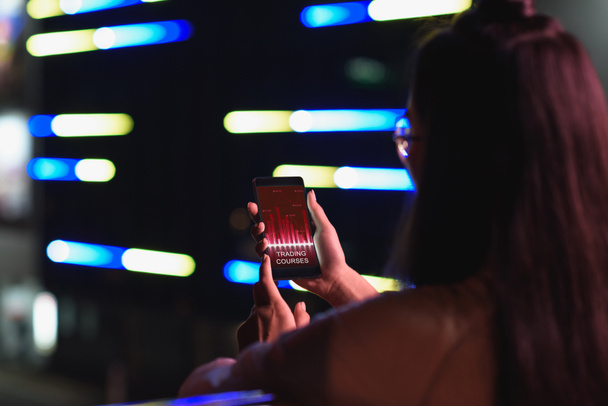 вид ззаду дівчини, що тримає смартфон із приладом для торгівлі на вулиці з неоновим світлом ввечері, місто майбутньої концепції
 - Фото, зображення