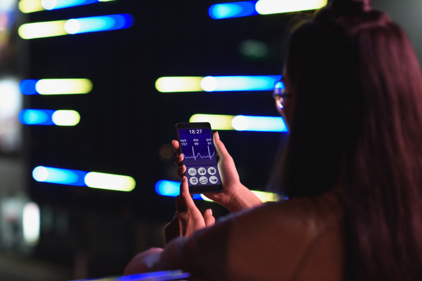 πίσω όψη του κορίτσι κρατώντας smartphone με ιατρική συσκευή στο δρόμο με φως νέον στο βράδυ, πόλη των μελλοντικών έννοια - Φωτογραφία, εικόνα