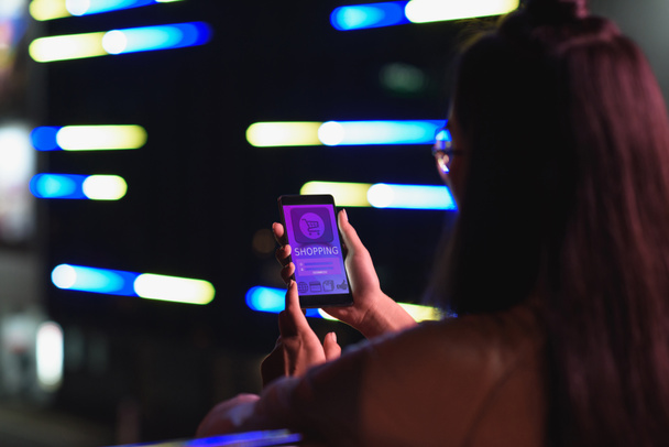 girl gazdaság smartphone-val bevásárló utcán neon fénnyel készülék este, a város jövőbeni koncepció hátulnézete - Fotó, kép