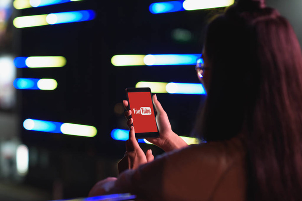 Achteraanzicht van meisje smartphone met geladen youtube pagina te houden op straat met neon licht in de avond, stad van de toekomst concept - Foto, afbeelding