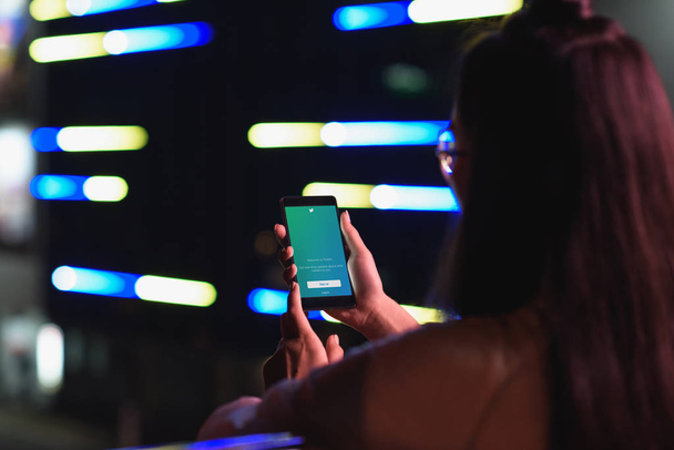 Achteraanzicht van meisje smartphone met geladen twitter-pagina houden op straat met neon licht in de avond, stad van de toekomst concept - Foto, afbeelding