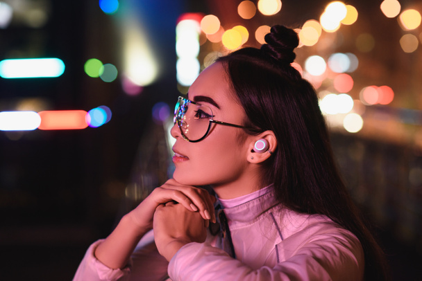 vista laterale di elegante ragazza asiatica in occhiali e auricolare wireless guardando lontano sulla strada con luce al neon in serata, città del concetto futuro
 - Foto, immagini