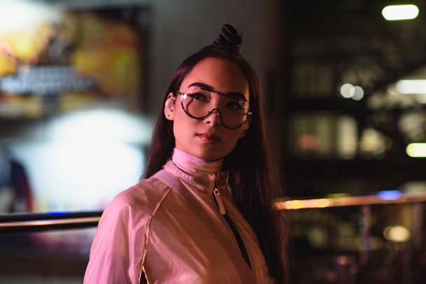メガネと無線イヤホンがネオンの光の通りに夕方に、未来の都市離れて見てスタイリッシュなアジアの女の子の肖像画 - 写真・画像