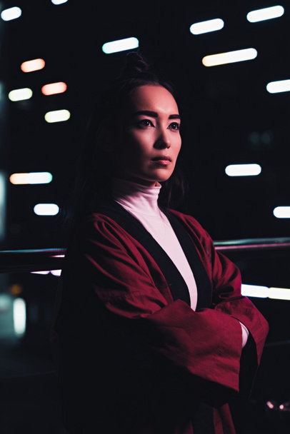 attraente ragazza asiatica in kimono bordeaux in piedi con le braccia incrociate sulla strada con luce al neon in serata, città del concetto futuro
 - Foto, immagini