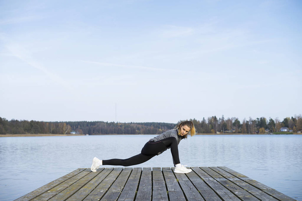 Young woman in sportswear stretching on pier near river  - Фото, изображение