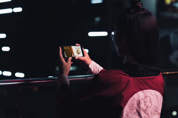 вид ззаду дівчини, що тримає смартфон з квитками онлайн-прилад на вулиці з неоновим світлом ввечері, місто майбутньої концепції
 - Фото, зображення