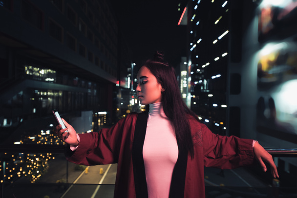 attractive asian girl leaning on railing and using smartphone on street with neon light in evening, city of future concept - Foto, Imagen