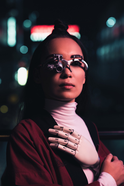 attractive asian girl with futuristic arm and eye prosthesis looking at camera on street in evening, city of future concept - Photo, Image