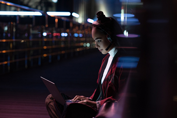attractive asian girl in kimono sitting and using laptop with headphones on street with neon light, city of future concept - Photo, Image