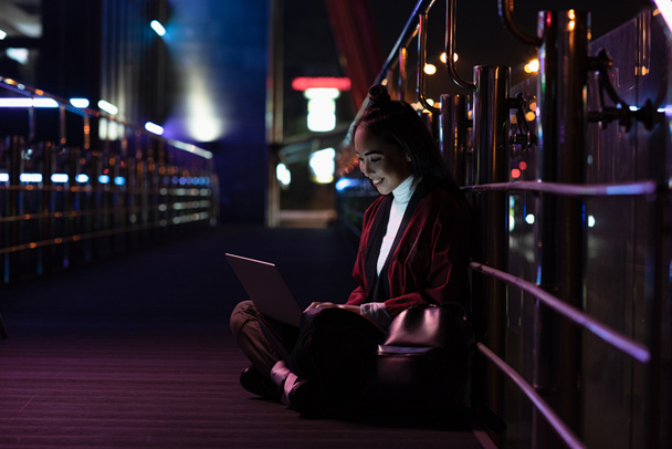 sonriente atractiva chica asiática en kimono sentado y usando portátil en la calle con luz de neón, ciudad de concepto futuro
 - Foto, Imagen