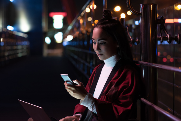 vonzó ázsiai lány ül, és a smartphone utcán neon fénnyel, város a jövőbeni koncepció laptop segítségével kimonó - Fotó, kép